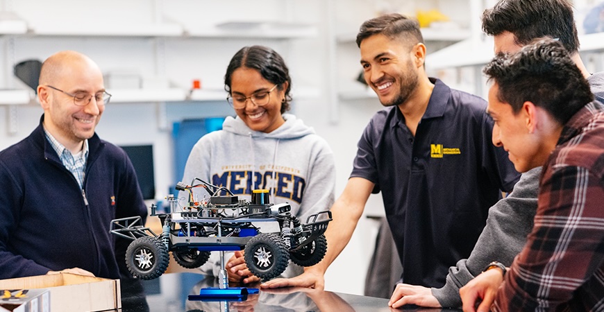 Depicted are five people looking at a robotic model car. 